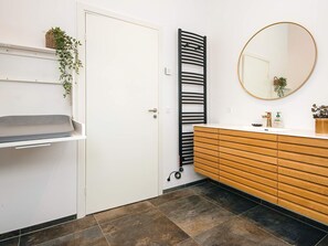 A bathroom with a white door, a towel rack, a round mirror, a wooden vanity, and a tiled floor.