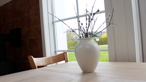 A white vase with branches and leaves on a table by a window.