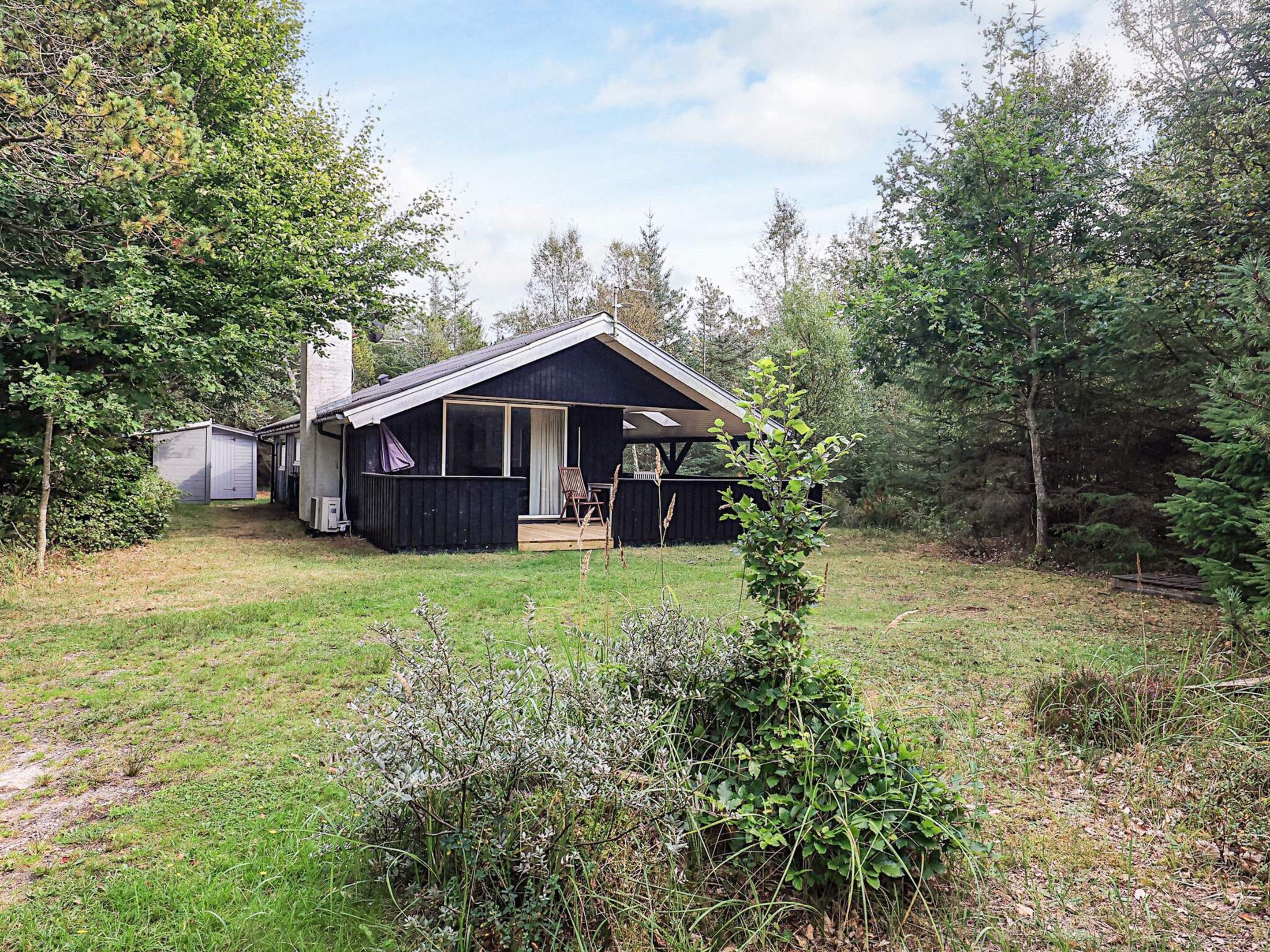 Une cabane avec un porche, un espace de détente extérieur abrité et un petit abri de jardin, le tout situé dans un cadre boisé.
