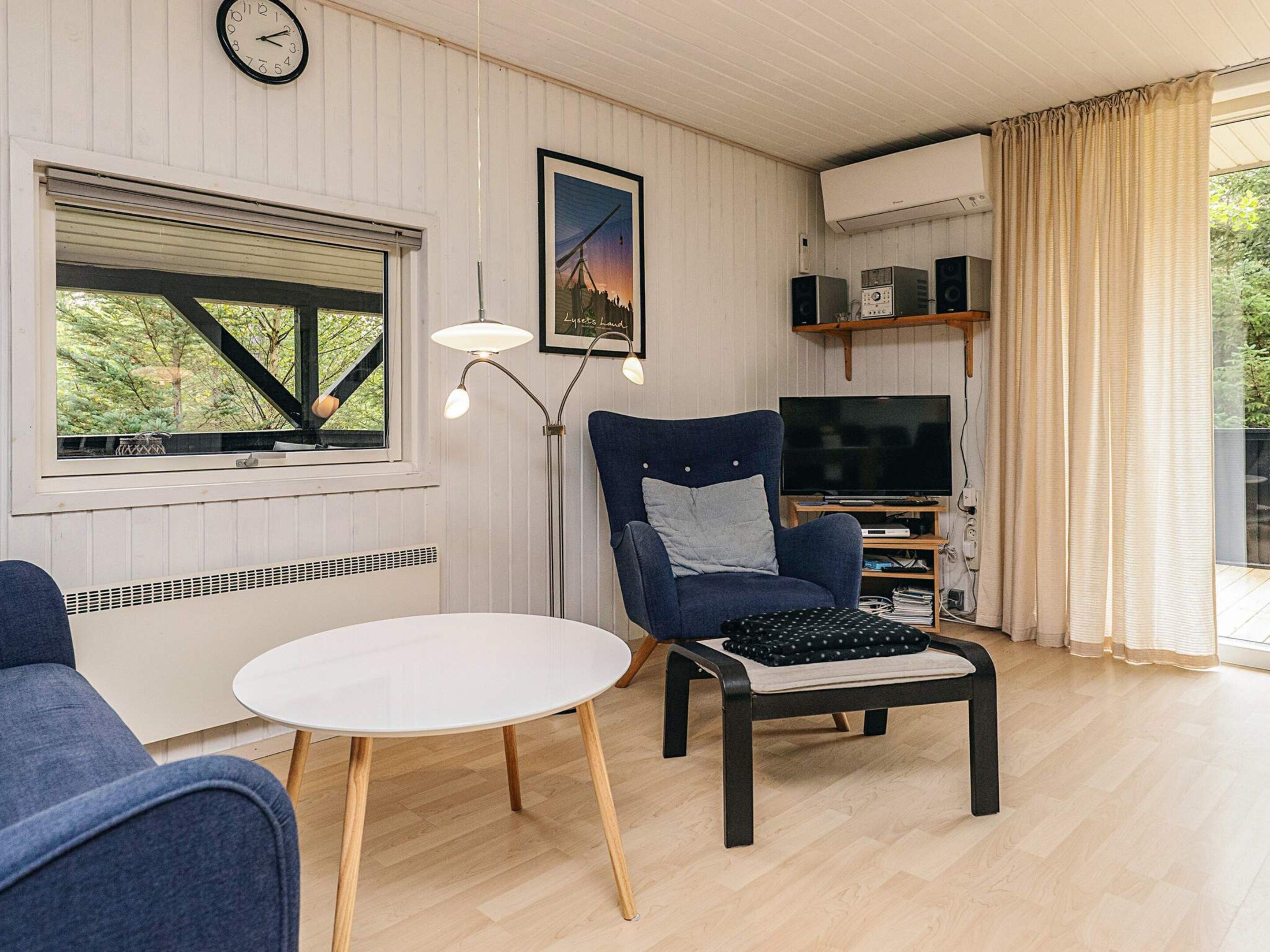 Una sala de estar con televisor, una silla, una mesa, un reloj y una ventana con persianas.