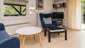 Un salon avec un téléviseur, un fauteuil, une table, une horloge et une fenêtre avec des stores.