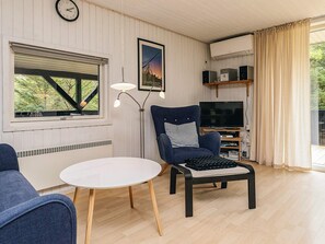 Un salon avec un téléviseur, un fauteuil, une table, une horloge et une fenêtre avec des stores.