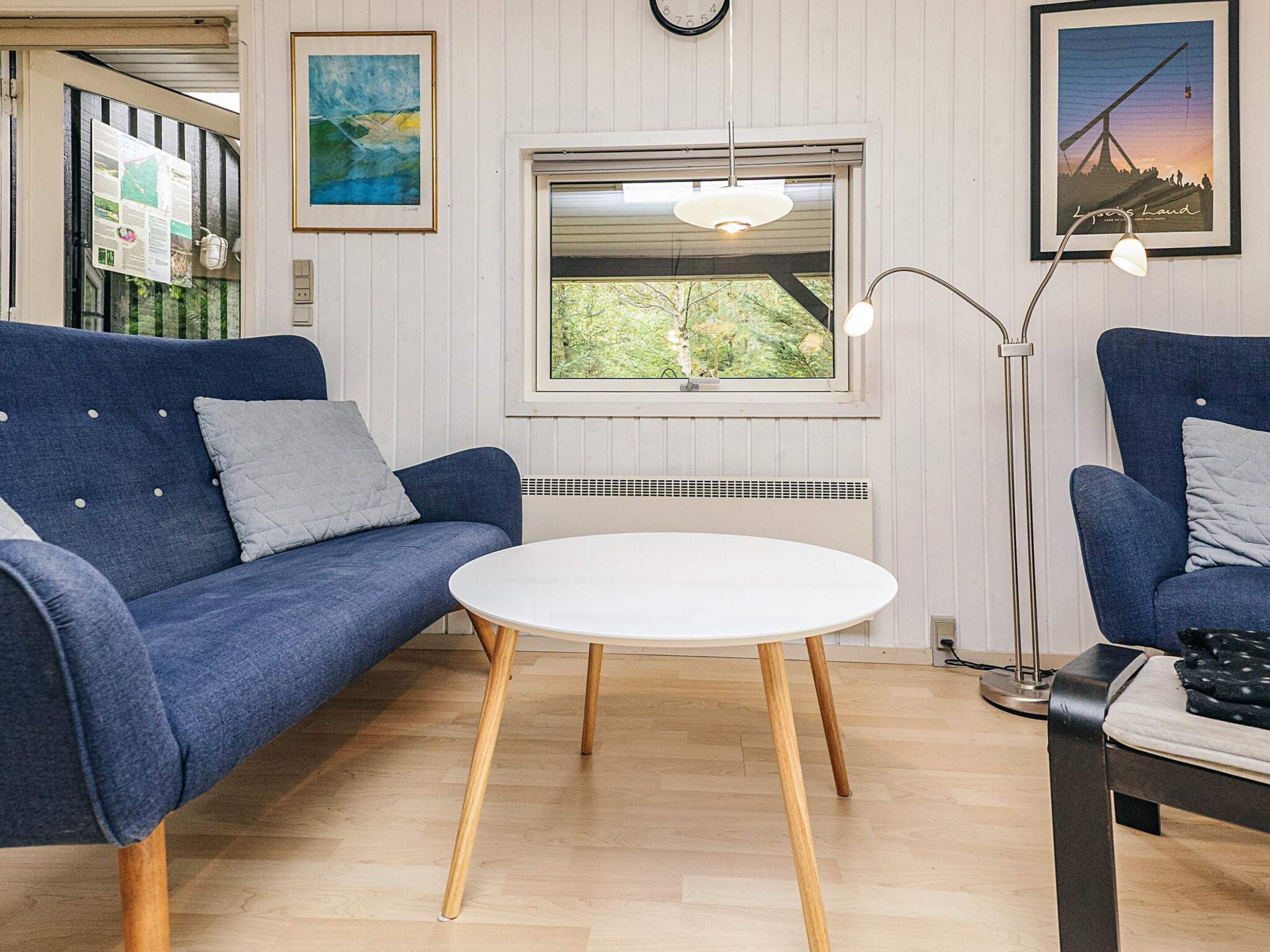 A room with a blue sofa, a white table, a floor lamp, and a window with a view of trees.