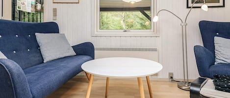 Une pièce avec un canapé bleu, une table blanche, un lampadaire et une fenêtre donnant sur des arbres.