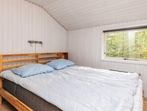 Une chambre avec un lit en bois, des murs blancs, une fenêtre donnant sur des arbres et un plafond avec des poutres en bois.