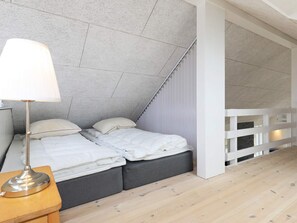 A bedroom with a sloped ceiling, a bed with grey bedding, a bedside table with a lamp, and a white railing. - 5 Star Holiday Home in Lokken (Lokken)