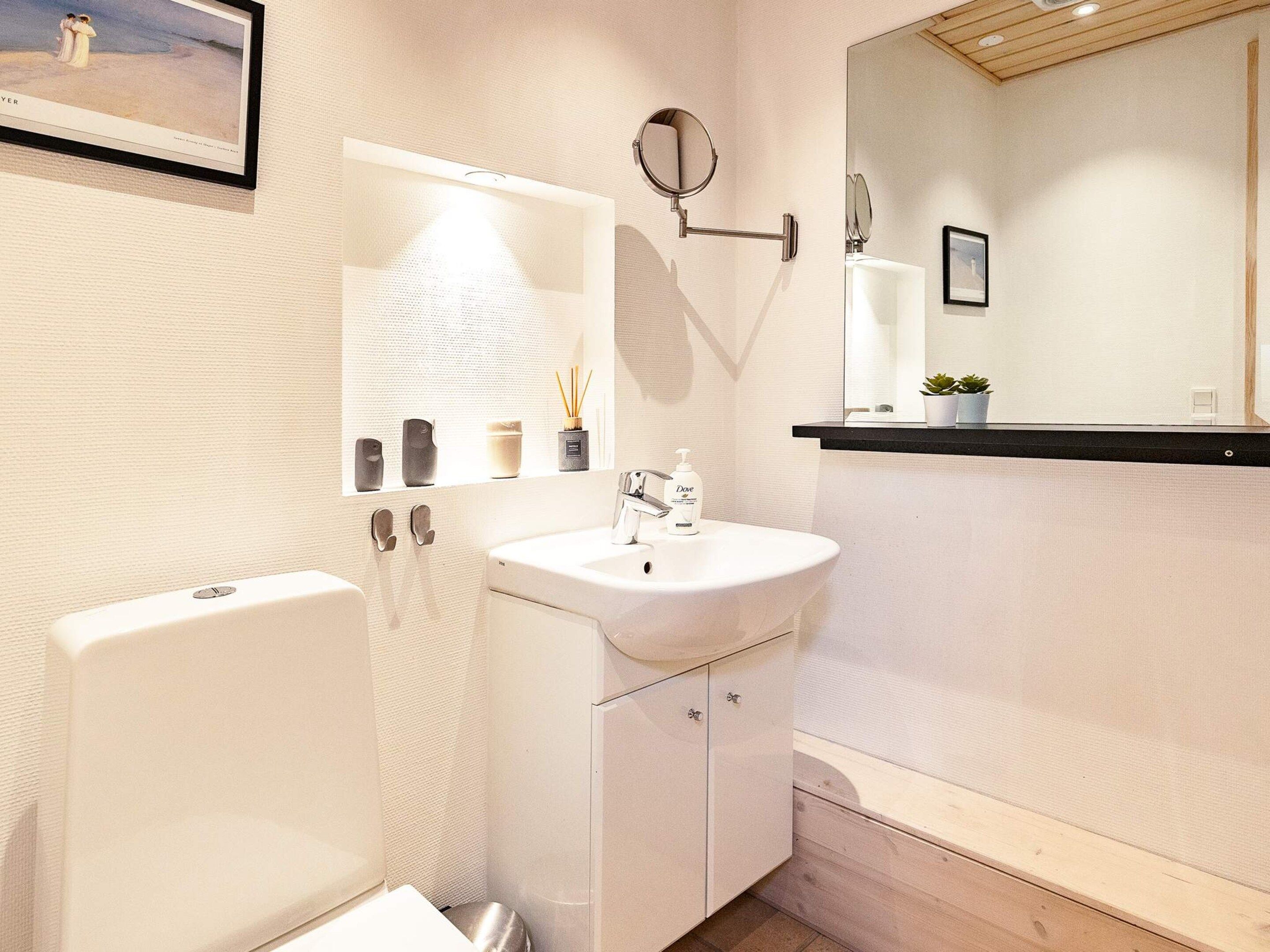 A bathroom with a white sink, a mirror, a toilet, and a shelf with toiletries.