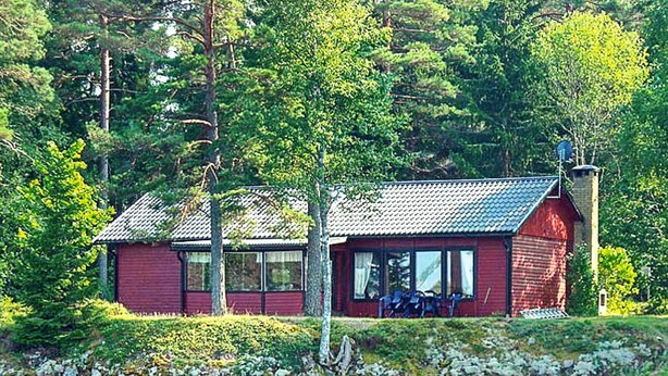 A red cabin by a lake with a dock, surrounded by trees. - 6 Person Holiday Home in Vena (Hultsfred)