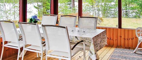 A covered outdoor dining area with a table and chairs, large windows, and a view of trees.
