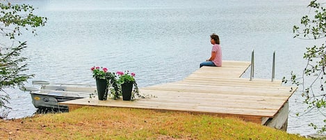 Un muelle de madera se adentra en un lago tranquilo, con una persona sentada y plantas en macetas cerca.
