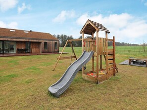 A wooden play structure with a slide and climbing elements, set in a grassy yard with a house in the background.