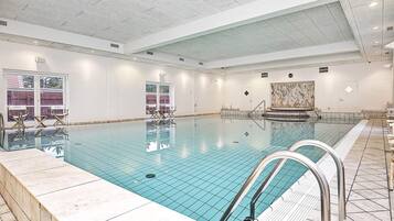 En stor indendørs swimmingpool med krystalklart vand, omgivet af hvide vægge og flisegulv.
