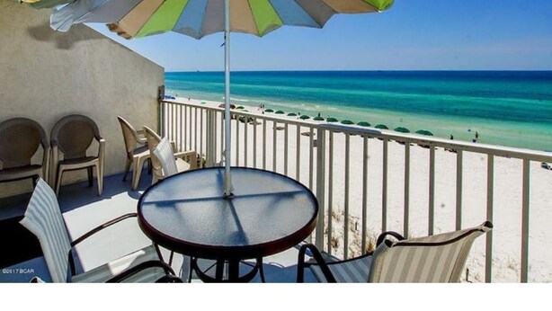 A balcony with a table and chairs, overlooking a beach with clear turquoise water and a white sandy shore. - BEACHfront 2-bedroom condo! (Panama City Beach)