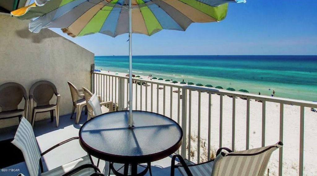 A balcony with a table and chairs, overlooking a beach with clear turquoise water and a white sandy shore.