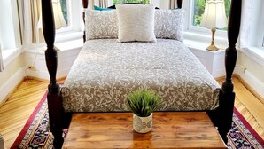 Ein Bett mit Baldachin, eine blumige Tagesdecke, ein kleiner Topf mit Pflanze auf einer Holzbank und Blick ins Grüne durch große Fenster.