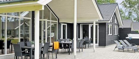 A covered outdoor patio with tables and chairs, a pool area with lounge chairs, and a building with a dark exterior and white trim.