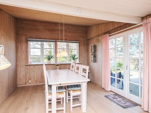 Un interior de madera con una mesa de comedor, sillas, una lámpara colgante y ventanas grandes con cortinas.