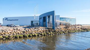 A modern building with a large sign reading 'Kattegatcentrum,' a parking area, and a rocky shoreline.