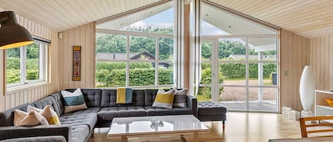 A modern living room with a sectional sofa, a glass coffee table, and large windows offering a view of greenery and buildings.