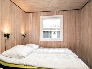 A bedroom with a wooden paneled wall, a single bed with white bedding, two wall-mounted lights, and a window with a view of a building outside.
