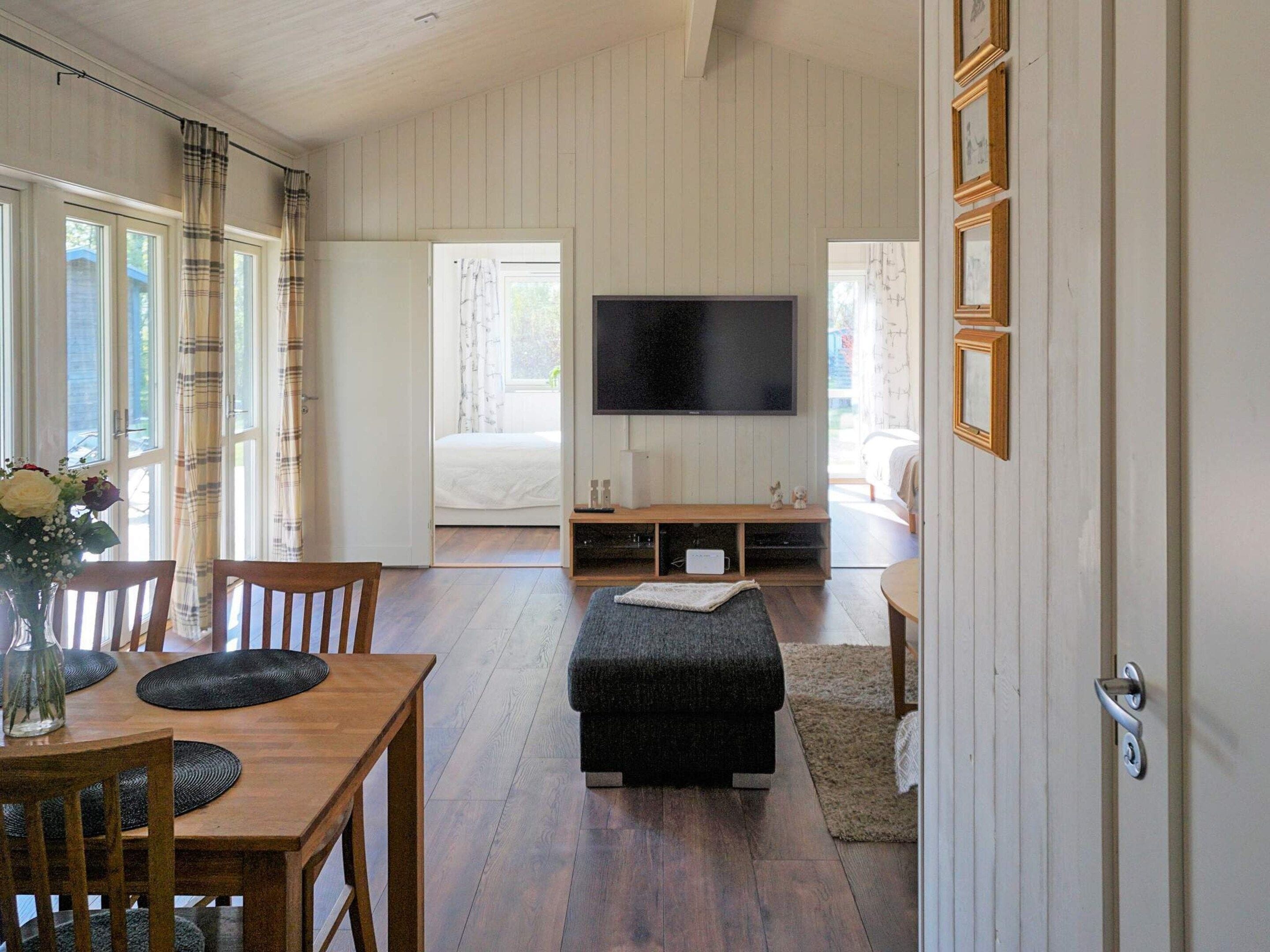 A room with a wooden dining table, chairs, a black ottoman, a flat-screen TV mounted on the wall, and a bed with a white headboard.