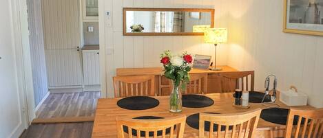 A dining area with a wooden table and chairs, a vase with flowers, a lamp, and a framed picture on the wall.