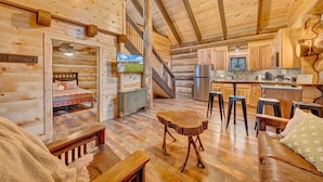 Interior de una cabaña de madera con una sala de estar acogedora, una cocina y una escalera que sube a un dormitorio estilo altillo.