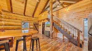 Interior de una cabaña de troncos con una mesa de comedor de madera, taburetes de bar, una escalera y una zona de cocina.