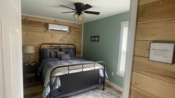 A bedroom with a bed, a ceiling fan, a wall-mounted air conditioner, a bedside table with a lamp, and a framed quote on the wall.
