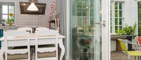 A dining area with a white table and chairs, a hanging pendant light, and a view through a glass door to an outdoor patio with chairs.