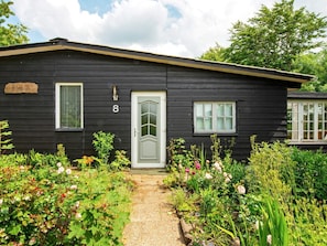 A black wooden house with a white door, number 8, and a small garden with various plants and flowers. - 4 Person Holiday Home in Skaerbaek (Skaerbaek)