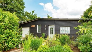 Une maison noire avec une porte et des fenêtres blanches, entourée d’une végétation luxuriante et de plantes en fleurs.