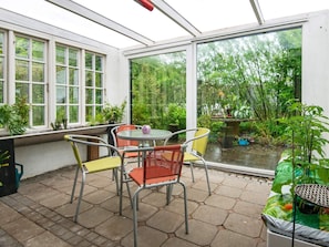 A conservatory with a glass roof, a round table, and chairs, surrounded by potted plants and a view of greenery. - 4 Person Holiday Home in Skaerbaek-by Traum (Skaerbaek)