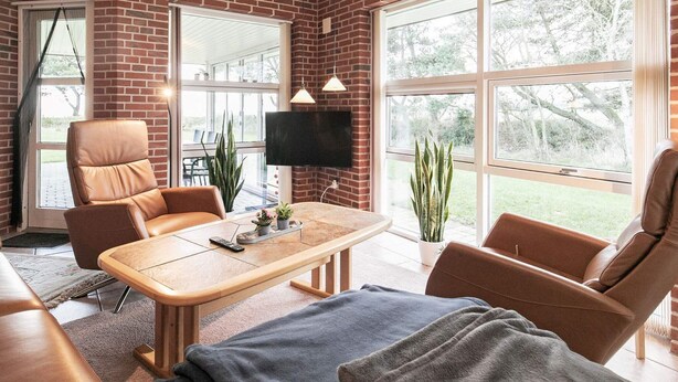 A modern living room with a brick wall, large windows, a flat-screen TV, leather chairs, and a wooden coffee table. - 6 Person Holiday Home in Blavand (Blavand)