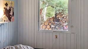 Une pièce avec un lit, une fenêtre donnant sur un arbre et une pile de bois de chauffage, ainsi qu’un luminaire fixé au mur.