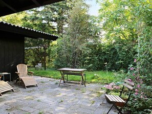 An outdoor patio area with a wooden table and chairs, surrounded by lush greenery and flowering plants.