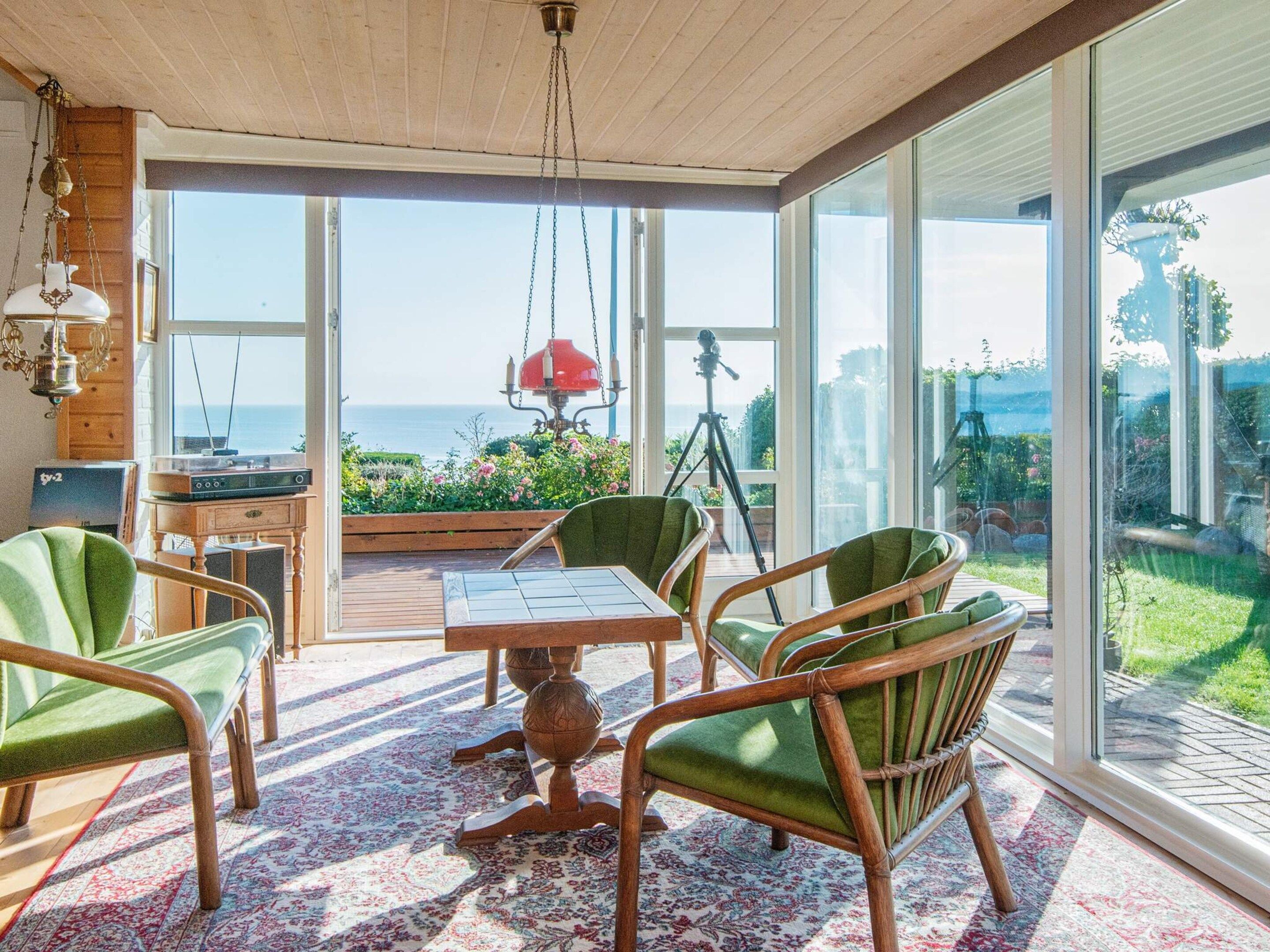 Une pièce dotée d’une grande porte vitrée donnant sur un balcon avec vue sur la mer. On y trouve une table avec des chaises, un luminaire suspendu et un trépied à l’arrière-plan.