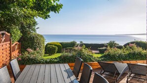 Un patio al aire libre con una mesa de madera y sillas de mimbre, con vistas a un jardín ajardinado y al mar.