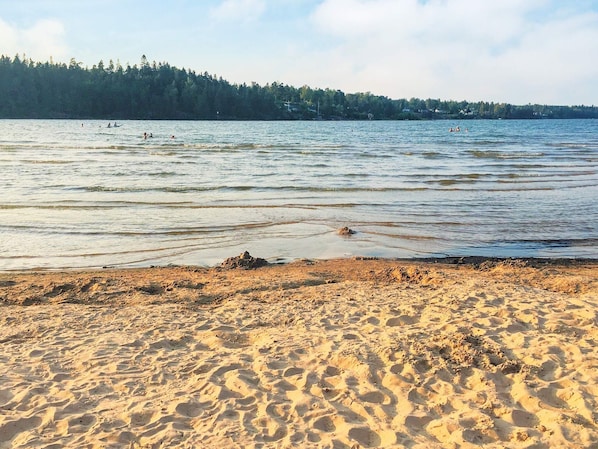 Una playa arenosa con huellas que van hacia el agua, un lago tranquilo y una colina boscosa al fondo.