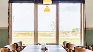 Une pièce avec une grande table et des chaises, une fenêtre donnant sur la plage et une lampe suspendue.