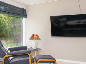 A modern living room with a flat-screen TV mounted on the wall, a grey sofa, a small round table with a decorative lamp, and a view of greenery outside. - 6 Person Holiday Home in Laeso (Læsø)