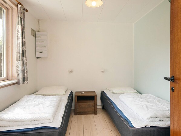A small, single-room accommodation with two beds, a wooden nightstand, a window with curtains, and a wall-mounted air conditioning unit.