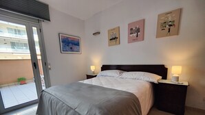 A neatly made bed with a grey bedspread, two bedside tables with lamps, and three wall art pieces depicting New York, Paris, and London.