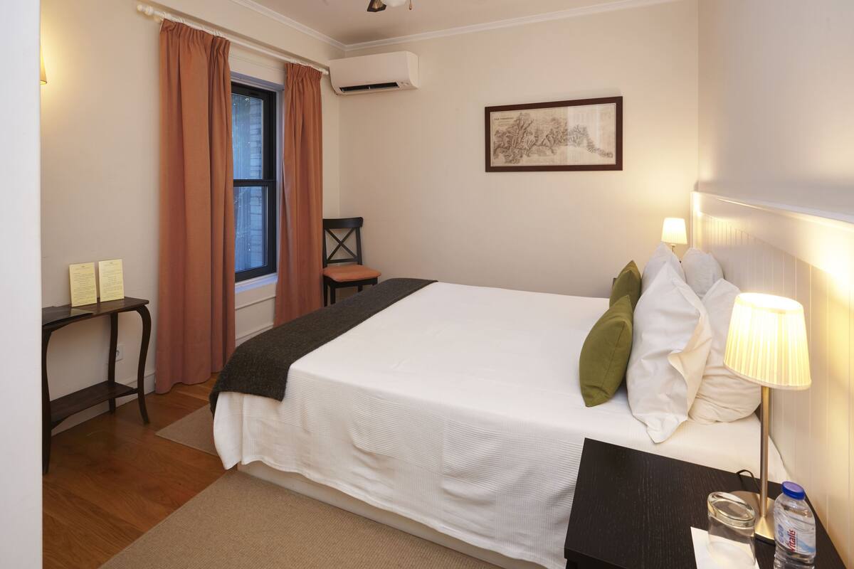 A hotel room with a bed, a desk, a chair, a window with curtains, an air conditioner, and a wall-mounted lamp.