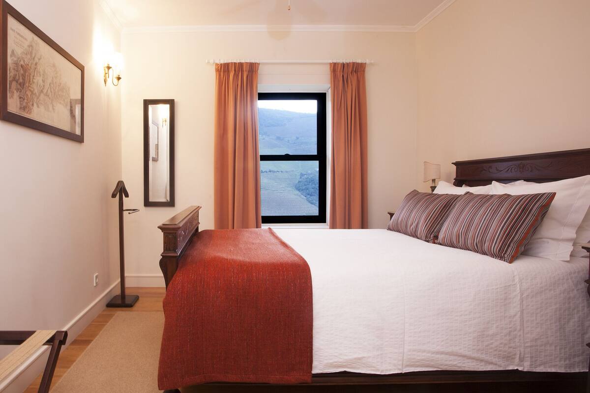 A bedroom with a large bed, a window with curtains, a bedside table, and a lamp.