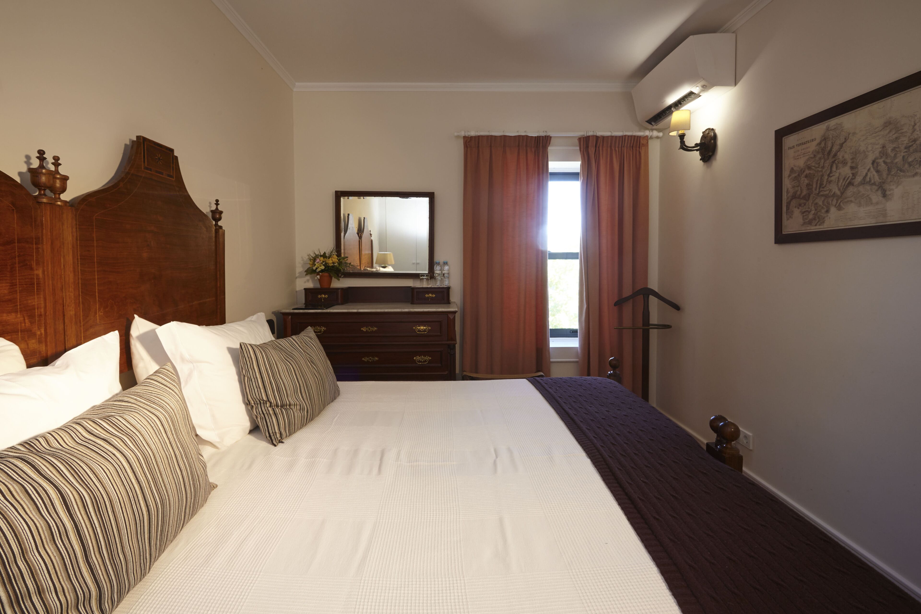 A neatly made bed with white and striped bedding, a wooden headboard, a dresser with a mirror, and a framed picture on the wall.