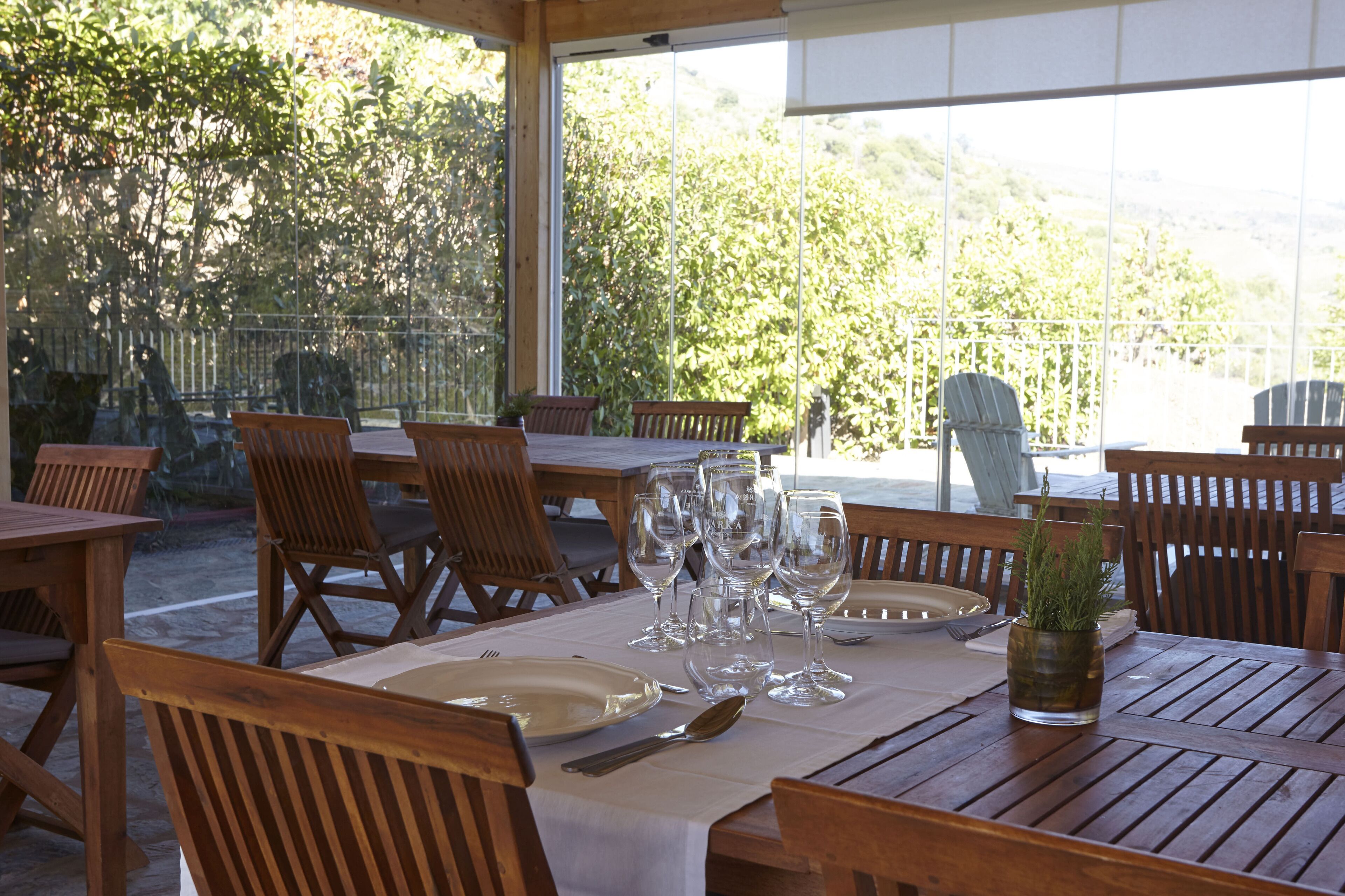 Un espacio de comedor al aire libre con mesas y sillas de madera, dispuesto con platos, cubiertos y copas de vino.