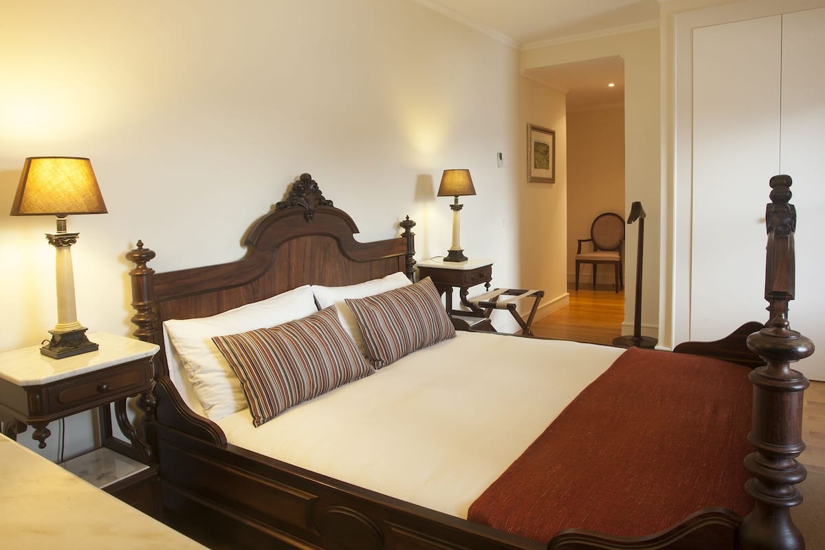 A neatly made bed with a wooden headboard, two bedside tables with lamps, a chair, and a door in the background.