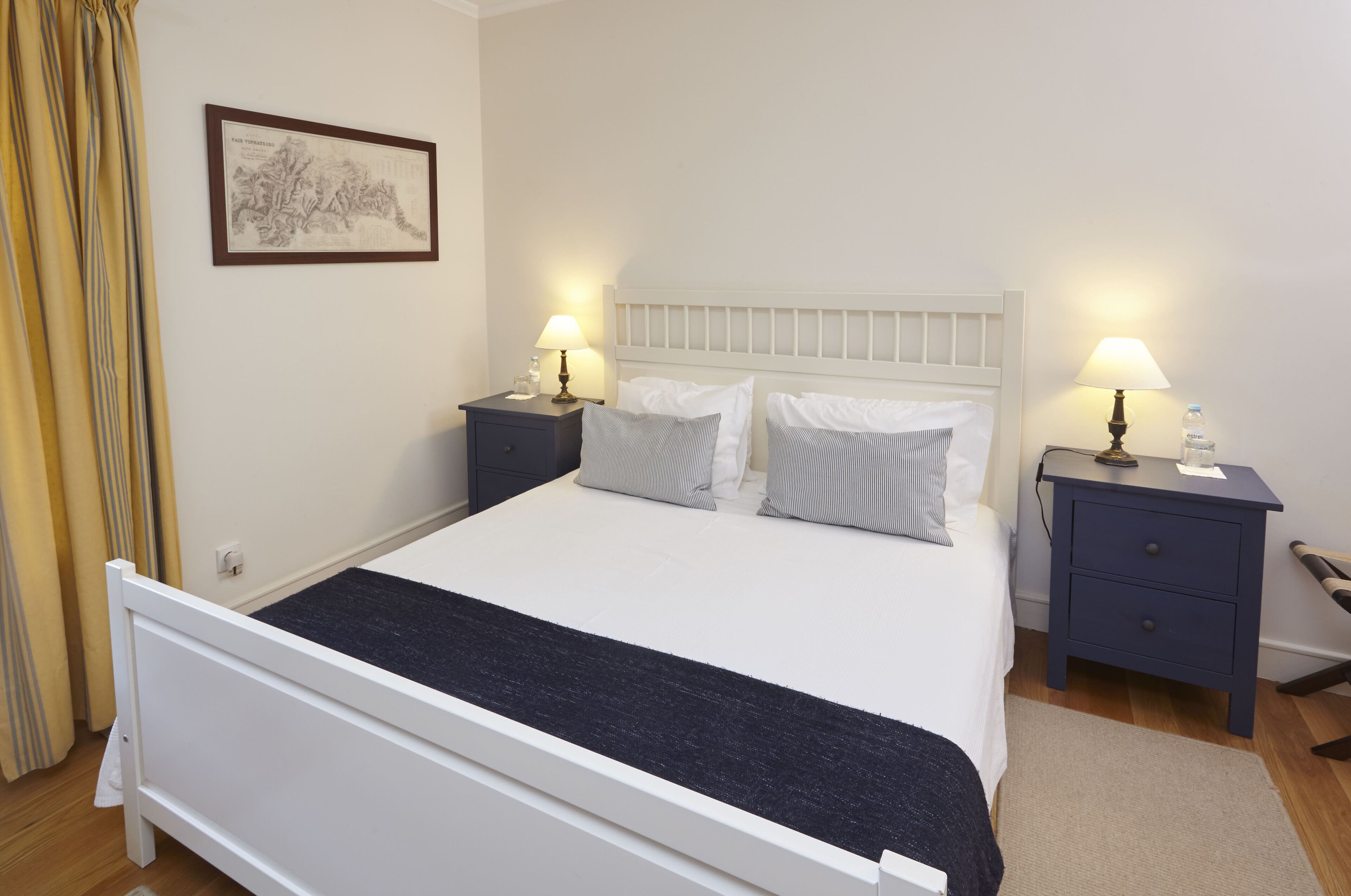A neatly arranged bedroom with a white bed, two bedside tables with lamps, a framed picture on the wall, and a curtain on the left.