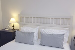 Una cama bien tendida con ropa de cama blanca y almohadas a rayas, una mesita de noche con una lámpara y una mesita auxiliar con una botella y un vaso.